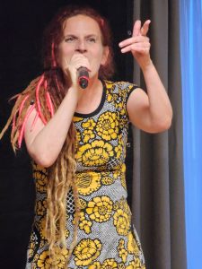 Mikaela Maniskkalalla on päällään graaffinen mekko jossa on keltaisia kukkia tummalla pohjalla. Ja hän osoittaa sormellaan yleisöön laulun sanojen mukaan.